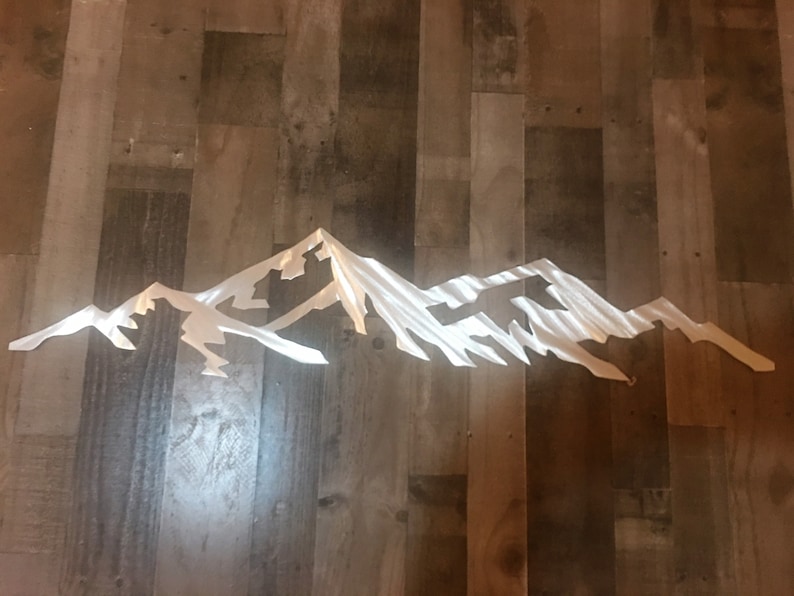 May include: A silver metal mountain range wall art sculpture. The sculpture is made of thin metal and has a brushed finish. The mountains are in a horizontal line and are all different sizes. The sculpture is perfect for adding a modern touch to any room.