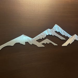 May include: A silver-colored metal wall art piece featuring a mountain range silhouette. The design consists of three distinct peaks with a brushed metallic finish that reflects light, creating a textured appearance across the surface of the metal.
