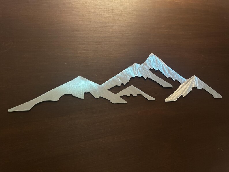 May include: A silver-colored metal wall art piece featuring a mountain range silhouette. The design consists of three distinct peaks with a brushed metallic finish that reflects light, creating a textured appearance across the surface of the metal.