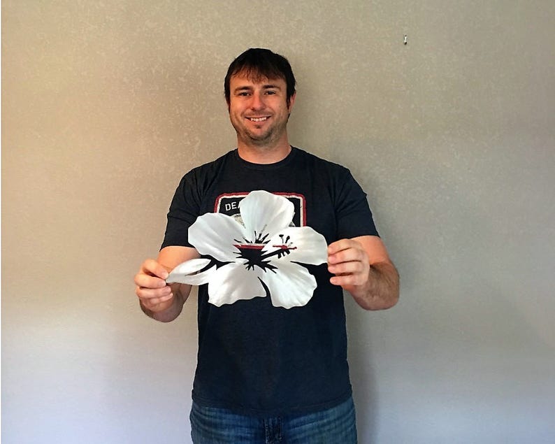 Puede incluir: Una persona sosteniendo un recorte de flor de hibisco de metal blanco. La flor mide aproximadamente 30 cm de ancho y tiene un centro rojo.