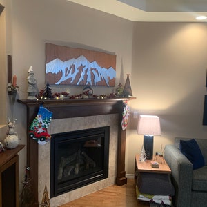 May include: A cozy living room with a fireplace decorated for Christmas. A wooden mountain art piece hangs above the fireplace. Christmas stockings, a snowman figurine, and a small Christmas tree are on the mantel. A lamp and side table are next to a gray sofa.