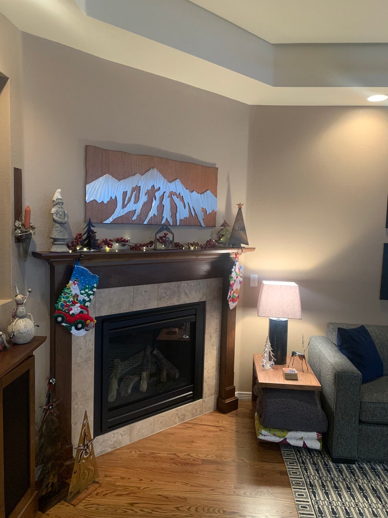 May include: A cozy living room with a fireplace decorated for Christmas. A wooden mountain art piece hangs above the fireplace. Christmas stockings, a snowman figurine, and a small Christmas tree are on the mantel. A lamp and side table are next to a gray sofa.