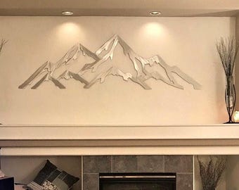 May include: A silver-toned metal wall art piece depicting a mountain range with jagged peaks and layered slopes. The sculpture features a reflective, smooth surface and is mounted horizontally on a flat, light-colored wall above a fireplace mantel.