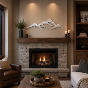 May include: A silver-toned metal mountain range wall art piece mounted above a stone fireplace. The sculpture features layered, jagged peaks with a metallic finish, positioned centrally on a neutral-colored wall above a dark wooden mantelpiece.