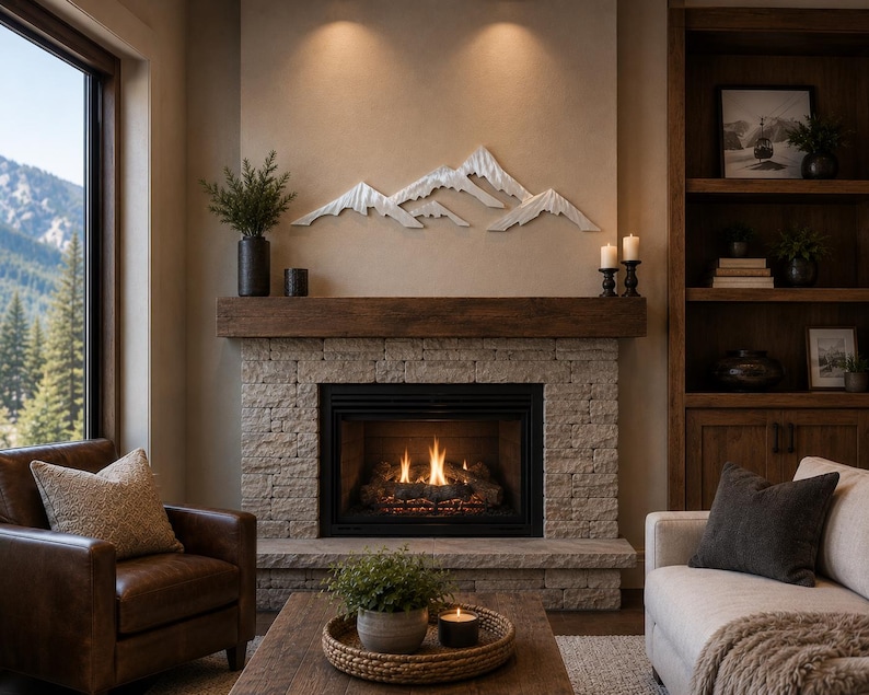 May include: A silver-toned metal mountain range wall art piece mounted above a stone fireplace. The sculpture features layered, jagged peaks with a metallic finish, positioned centrally on a neutral-colored wall above a dark wooden mantelpiece.