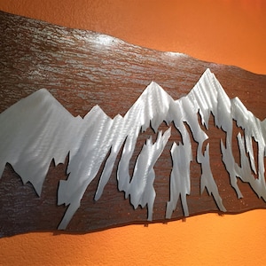 May include: Metal wall art depicting a mountain range. The mountains are cut out of silver-colored metal and set against a textured, brown background. The artwork is mounted on an orange wall.