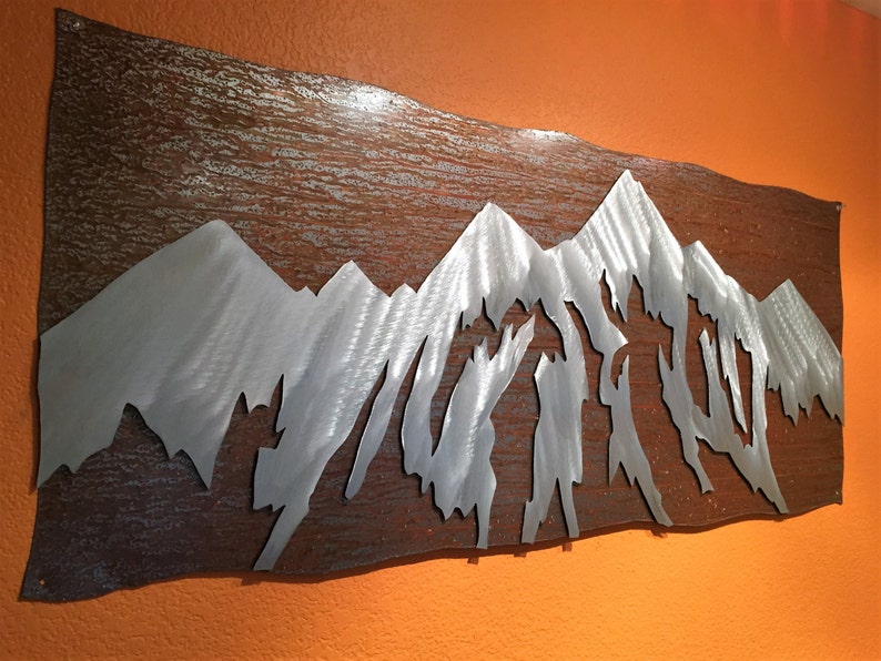 May include: Metal wall art depicting a mountain range. The mountains are cut out of silver-colored metal and set against a textured, brown background. The artwork is mounted on an orange wall.