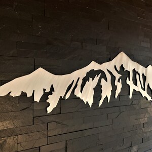 Pu&ograve; includere: Un decoro da parete in metallo bianco a forma di catena montuosa con una silhouette dettagliata di vette e valli. Il decoro &egrave; montato su una parete in mattoni neri.