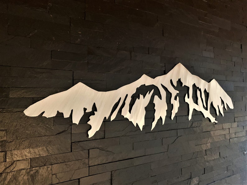 Pu&ograve; includere: Un decoro da parete in metallo bianco a forma di catena montuosa con una silhouette dettagliata di vette e valli. Il decoro &egrave; montato su una parete in mattoni neri.