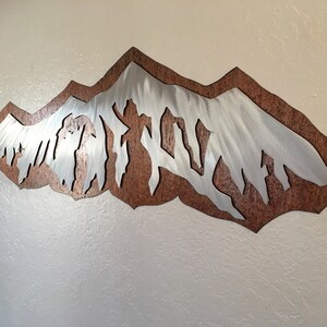 May include: A wall art piece featuring a silver-toned metal mountain range silhouette layered over a textured, rust-colored metal backing. The mountain peaks are jagged and angular, creating a sharp contrast against the dark, mottled brown background.