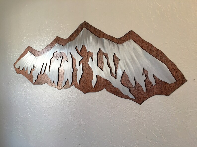May include: A wall art piece featuring a silver-toned metal mountain range silhouette layered over a textured, rust-colored metal backing. The mountain peaks are jagged and angular, creating a sharp contrast against the dark, mottled brown background.