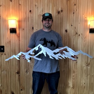 May include: A wide, silver-colored metal wall art piece featuring a jagged, multi-peaked mountain range silhouette. The design is crafted from a single sheet of metal with sharp, angular cutouts representing snow-capped peaks and rocky terrain.