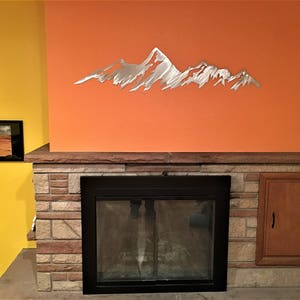 May include: A silver metal mountain range wall art decor is mounted on an orange wall above a fireplace. The fireplace is framed by a stone facade and a wooden cabinet. Two framed landscape photos hang on a yellow wall.