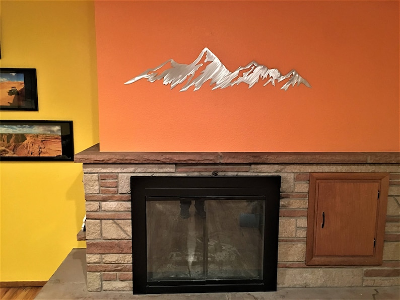 May include: A silver metal mountain range wall art decor is mounted on an orange wall above a fireplace. The fireplace is framed by a stone facade and a wooden cabinet. Two framed landscape photos hang on a yellow wall.