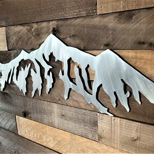 Pu&ograve; includere: Una silhouette di una catena montuosa in metallo, dipinta in finitura argentata, montata su una parete in pannelli di legno.