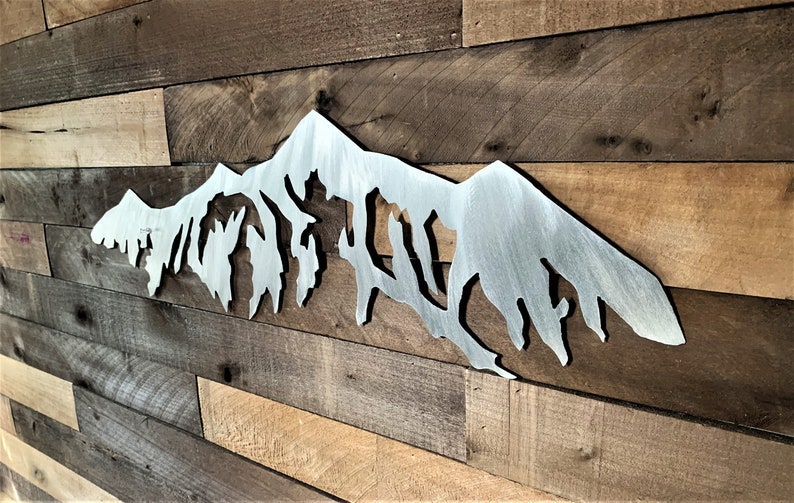 Pu&ograve; includere: Una silhouette di una catena montuosa in metallo, dipinta in finitura argentata, montata su una parete in pannelli di legno.