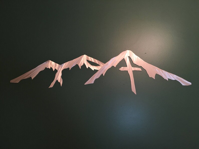 May include: A metal wall art sculpture depicting a mountain range with a cross on the highest peak. The sculpture is made of a brushed metal with a copper finish.