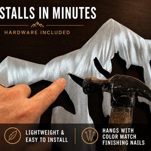 May include: A silver-colored metal mountain range wall art piece featuring a brushed finish. The text reads: INSTALLS IN MINUTES, HARDWARE INCLUDED, LIGHTWEIGHT & EASY TO INSTALL, and HANGS WITH COLOR MATCH FINISHING NAILS. A hammer is shown.
