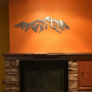 Pu&ograve; includere: Una scultura da parete in metallo a forma di catena montuosa, di colore argento, montata su una parete arancione sopra un camino in mattoni con un inserto per camino nero.