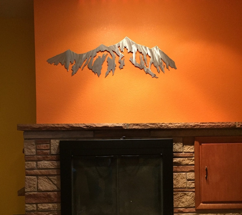 Pu&ograve; includere: Una scultura da parete in metallo a forma di catena montuosa, di colore argento, montata su una parete arancione sopra un camino in mattoni con un inserto per camino nero.