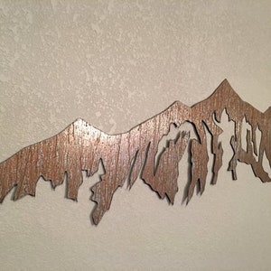 May include: A metal wall art sculpture of a mountain range with a rustic, weathered finish. The mountain peaks are cut out to create a silhouette effect.
