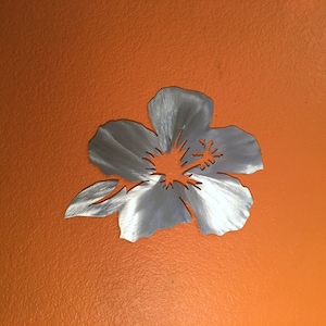 Puede incluir: Un recorte de flor de hibisco de metal plateado, con una superficie texturizada, sobre un fondo naranja brillante.