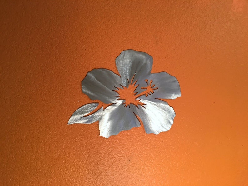 Puede incluir: Un recorte de flor de hibisco de metal plateado, con una superficie texturizada, sobre un fondo naranja brillante.