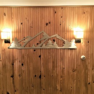 May include: A metal mountain range wall decor with trees, mounted on a wood panel wall with two light fixtures on either side.