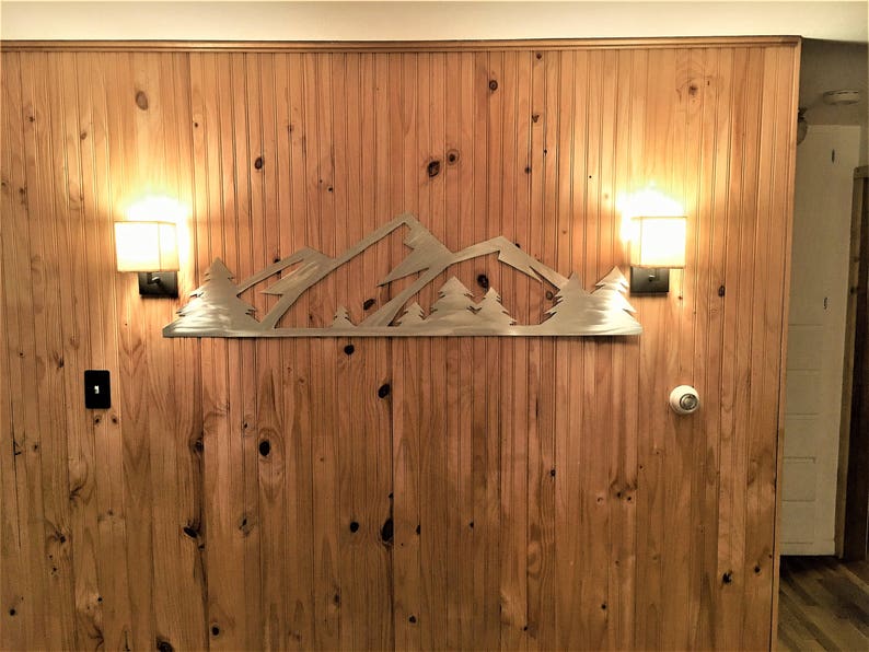 May include: A metal mountain range wall decor with trees, mounted on a wood panel wall with two light fixtures on either side.
