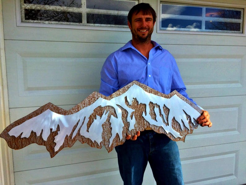 May include: A wide, decorative wall art piece shaped like a mountain range. The design features a textured, brown, bark-like base layer with a smooth, reflective, silver-colored metal overlay that mimics the appearance of snow-capped peaks.