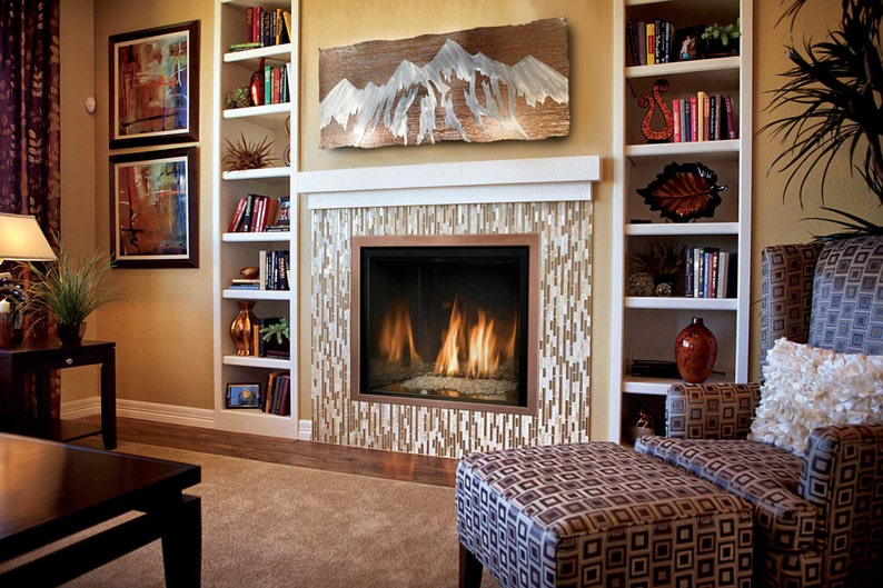May include: A cozy living room with a fireplace, bookshelves, and comfortable seating. The fireplace has a mosaic tile surround and a copper-colored frame. Above the fireplace is a wooden art piece depicting a mountain range.