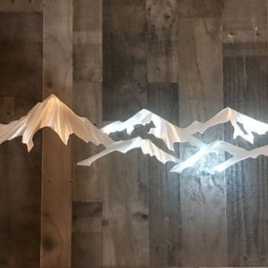 May include: A white mountain range wall art piece featuring layered, jagged peaks with a smooth finish. The design consists of multiple overlapping silhouettes that create a sense of depth and elevation, showcasing sharp, angular ridges and slopes.