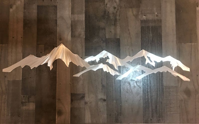 May include: A white mountain range wall art piece featuring layered, jagged peaks with a smooth finish. The design consists of multiple overlapping silhouettes that create a sense of depth and elevation, showcasing sharp, angular ridges and slopes.