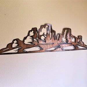 Puede incluir: Una pieza de arte mural de madera que representa una silueta de una cordillera. La obra de arte est&aacute; cortada de madera y te&ntilde;ida en un color marr&oacute;n c&aacute;lido, con &aacute;reas m&aacute;s claras que crean contraste. La pieza est&aacute; montada en una pared de color neutro.