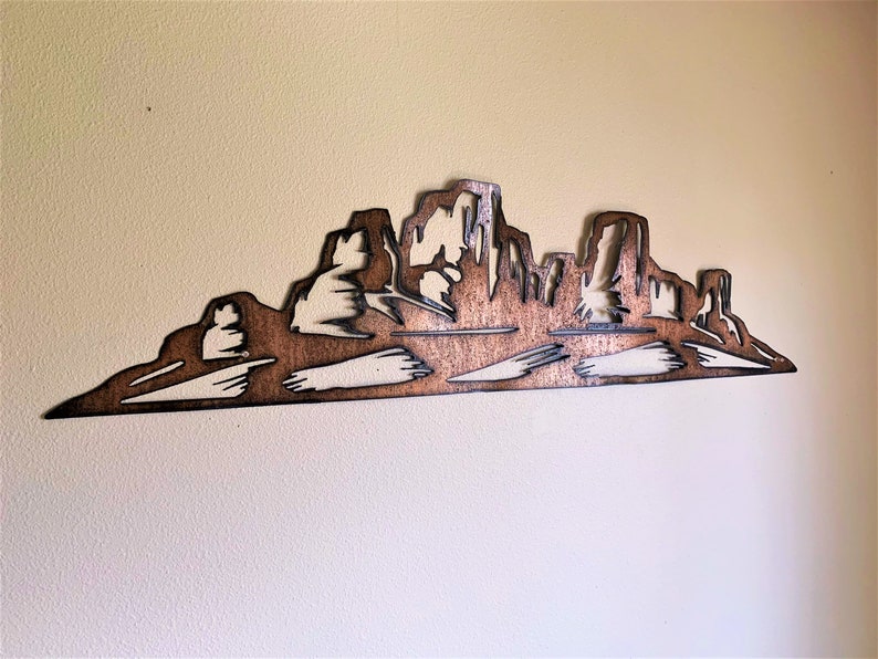 Puede incluir: Una pieza de arte mural de madera que representa una silueta de una cordillera. La obra de arte est&aacute; cortada de madera y te&ntilde;ida en un color marr&oacute;n c&aacute;lido, con &aacute;reas m&aacute;s claras que crean contraste. La pieza est&aacute; montada en una pared de color neutro.