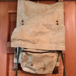 May include: A vintage canvas backpack with a brown leather zipper and green straps. The backpack is hanging on a wooden door.