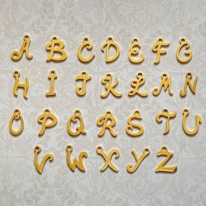 May include: Gold-colored metal alphabet letter charms. Each letter is individually crafted with a cursive font. The letters are arranged in alphabetical order.