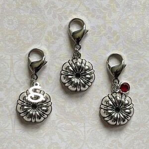 May include: Three silver-toned pumpkin charms with lobster claw clasps. One charm has the letter "S", another has a red gemstone, and the third is plain. These charms are suitable for jewelry making or seasonal crafts.