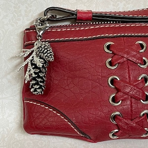 May include: A red leather wristlet with white stitching and a silver-tone pinecone charm. The wristlet features a zipper closure and a decorative lace-up design on the front. The wristlet has a wrist strap.