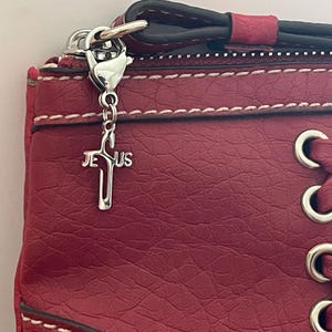 May include: Red handbag with white stitching and silver hardware. A silver cross charm with the word "JESUS" hangs from the zipper. The bag has a textured surface and a lace-up design on the side.