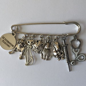 May include: A silver-toned safety pin brooch with charms. Charms include a dog, cat, guinea pig, rabbit, dog, syringe, and stethoscope. A round charm reads "VETERINARIAN ASSISTANT".