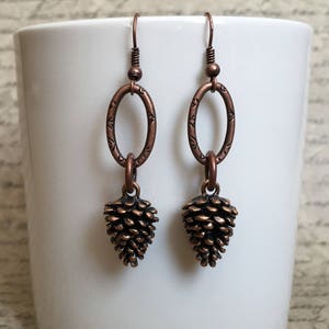 May include: A pair of copper-colored earrings. Each earring features a pinecone charm suspended from an oval-shaped link with etched detailing. The earrings hang from a simple hook.