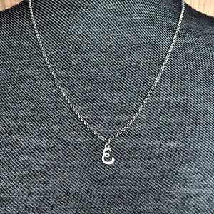 May include: A silver-colored necklace with a delicate chain and a small pendant shaped like the letter "E". The necklace is displayed on a dark gray mannequin, set against a wooden background.