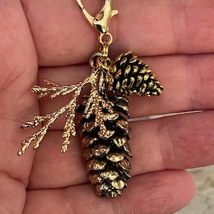May include: A gold-coloured charm featuring a detailed pinecone and sprig design. The charm includes a lobster clasp for easy attachment. The pinecone has a textured surface, and the sprig adds a natural element to the charm.