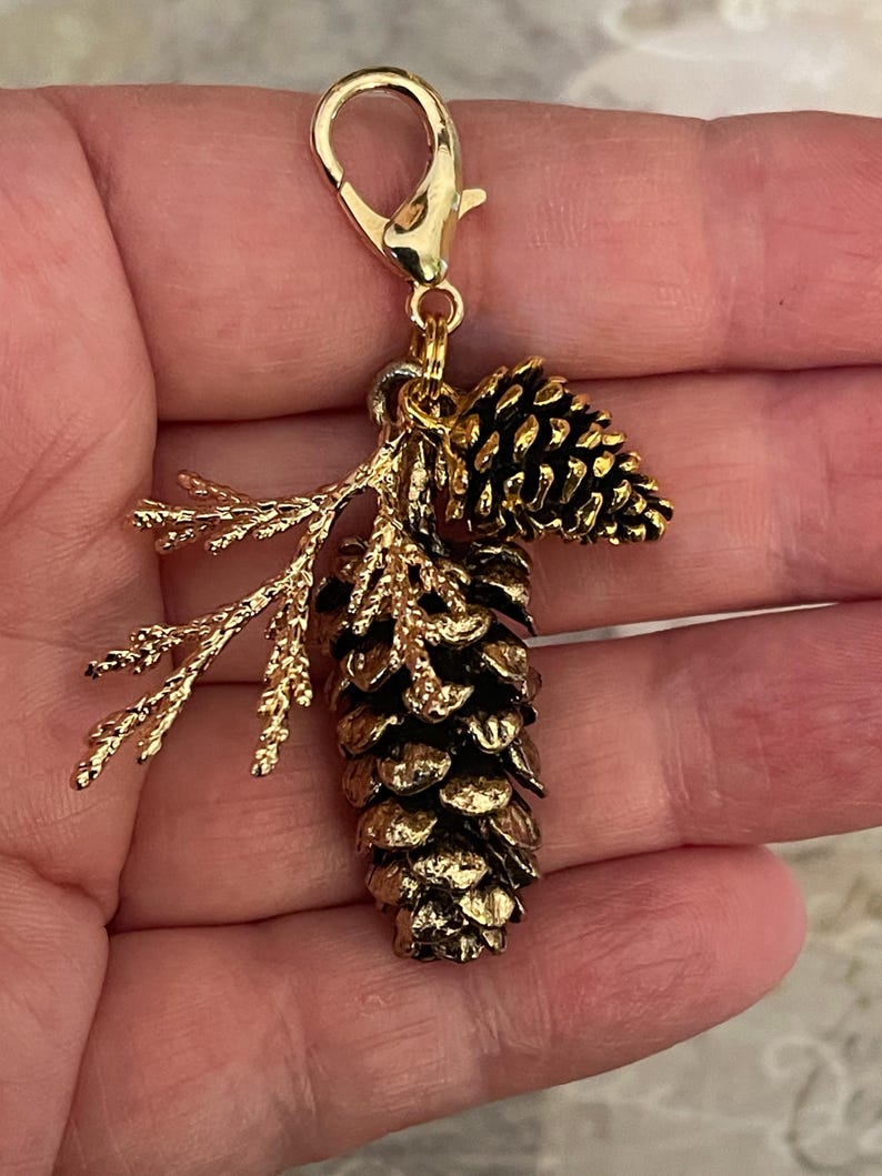 May include: A gold-coloured charm featuring a detailed pinecone and sprig design. The charm includes a lobster clasp for easy attachment. The pinecone has a textured surface, and the sprig adds a natural element to the charm.
