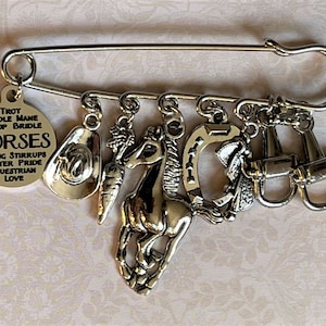 May include: Silver-toned safety pin with equestrian-themed charms. Charms include a cowboy hat, carrot, running horse, horseshoe, saddle, and riding equipment. A circular tag reads "HORSES" and related words. A unique accessory for horse lovers.