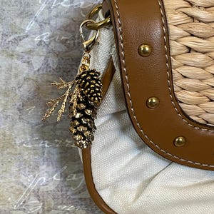 May include: A gold-coloured pine cone charm with a sprig of leaves hangs from a purse. The purse has a woven straw body, a brown leather strap with gold studs, and a white canvas interior. The charm is attached with a gold clasp.