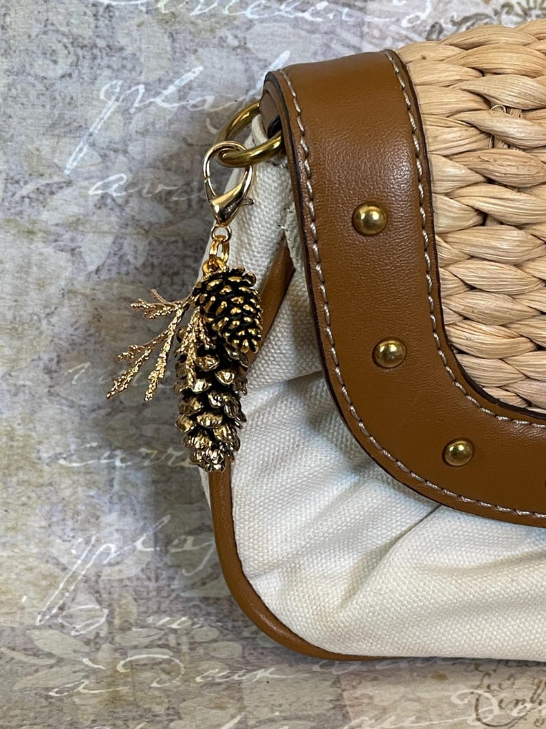 May include: A gold-coloured pine cone charm with a sprig of leaves hangs from a purse. The purse has a woven straw body, a brown leather strap with gold studs, and a white canvas interior. The charm is attached with a gold clasp.