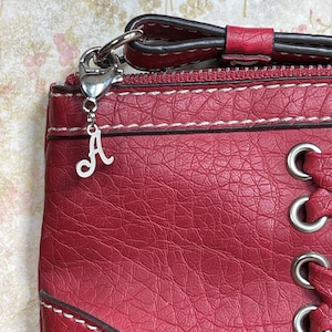 May include: A red leather purse with white stitching and silver hardware. A silver charm with the letter "A" hangs from the purse's zipper. The purse has a textured surface and a decorative lace-up design.