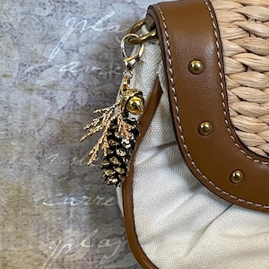 May include: A handbag with a woven straw body and brown leather trim. A gold-coloured charm shaped like a pinecone and sprig hangs from the bag's strap. The bag has a white canvas interior.